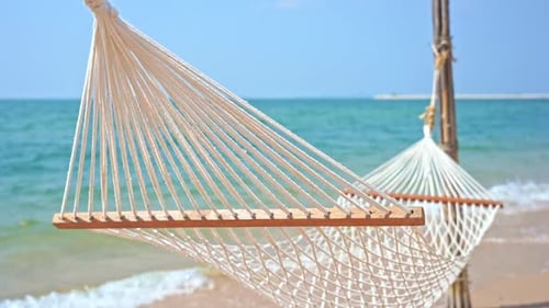 Static shot of an empty hammock slowly moving by the wind with calm waves reaching the shore from th