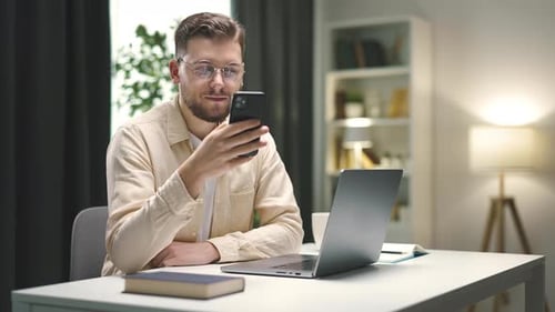 Man Looks at Smart Phone While at Desk