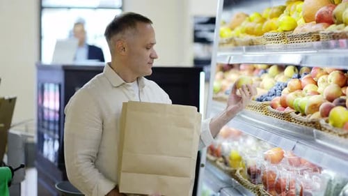 A Middleaged Shop Visitor Selects Apples From a Display Case and Puts Them Into a Paper Bag