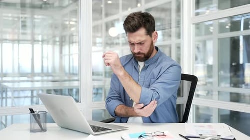 Businessman suffering from elbow pain while sitting at desk at workplace in business office. Upset