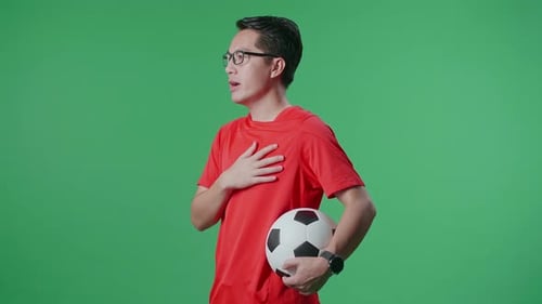 Side View Of Asian Man With A Ball Singing National Anthem While Cheering Soccer On Green Screen