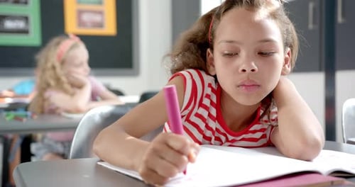 Biracial girl with light brown hair is focused on writing in a notebook in a classroom at school