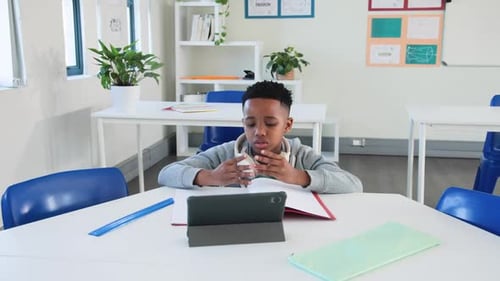 African preteen boy sitting at class table, adjusting phones, writing notes from tablet for study