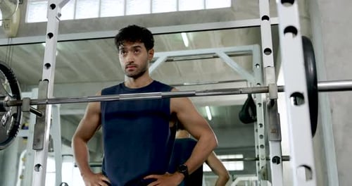 Young Asian man rests beside barbell rack in modern gym after shoulder workout, standing with hands