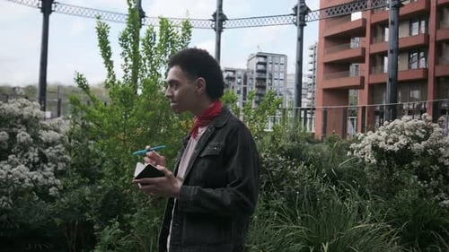 Joven periodista tomando notas al aire libre cerca de edificios modernos
