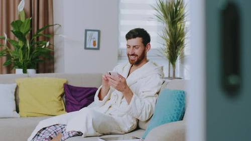 Handsome Young Man in Domestic Clothing Sitting on Couch Smiling Use Phone Feel Happy Stay at Home