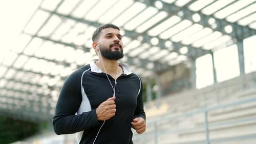 Adult bearded male runner in headphones jogging outside in urban city stadium. Fit handsome man