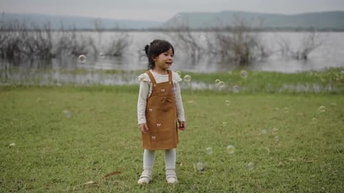 slow motion of happy toddler girl playing soap bubble in grass field near the river