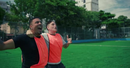 Latin and White friends joyfully celebrating goal on urban soccer field, cheering loudly, raising