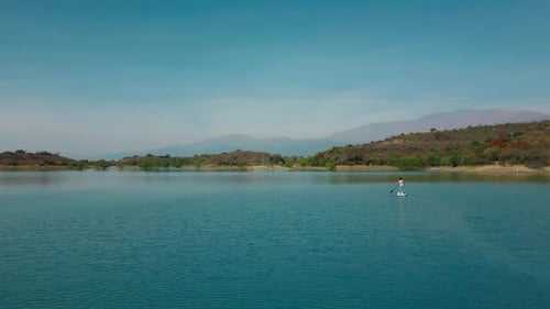 Aerial drone flying over a man doing stand up paddle in a blue water lake with mountains