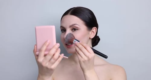 Beautiful woman applying face powder with brush on light grey background