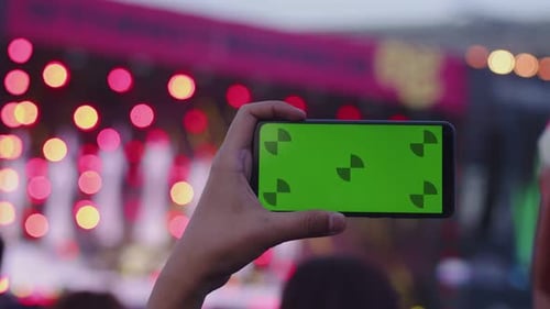 A Hand Holds a Smartphone with a Green Screen Chromakey and Shoots an Outdoor Concert the Lamps