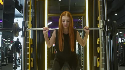 A Graceful and Athletic Redhead Performs Squats with a Barbell on Her Shoulders in the Gym