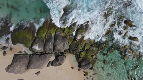 Waves of ocean crashing and flooding mossy rocks and sandy beach at shore during sunlight.