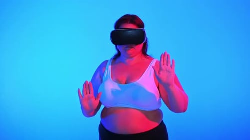 Woman with overweight in a tracksuit and VR headset posing in a studio with blue background and red