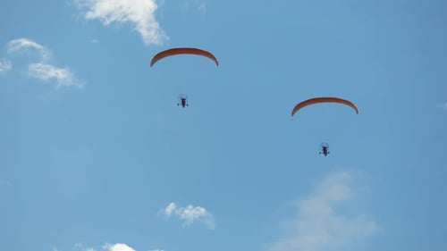 Two paramotor pilots operate powered paragliders with orange canopies, soaring high against vibrant