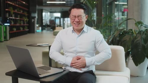 Disappointed Chinese Businessman Using Laptop and Feeling Sudden Stomach Ache in Office Lobby