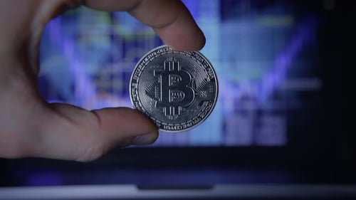 A Bitcoin Cryptocurrency Coin Held in Hand Set Against a Backdrop of Market Data