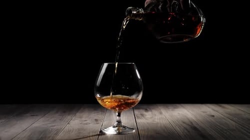 Conhaque de luxo. Mão derrama conhaque dourado de uma garrafa redonda em um copo na mesa de madeira
contra o fundo