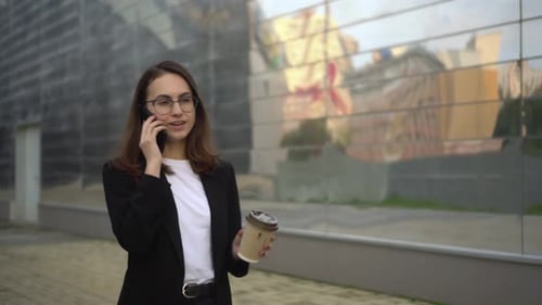 A Young Businesswoman is Talking on the Phone and Drinking Coffee Walking Around the Business Center