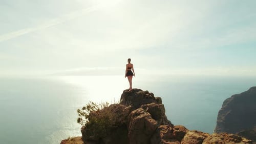 A woman stands on a rocky cliff overlooking the ocean under a bright sky. The wide landscape suggest