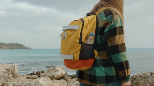 A woman in a stylish warm checkered shirt and an orange backpack strolls along the stones by the sea