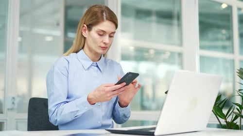 Mature businesswoman is using smartphone while sitting at workplace at desk in modern office. Woman