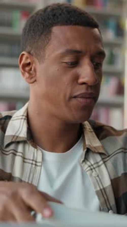 Focused African American Man Male Student in Library Putting on Glasses Reading Open Book Turning