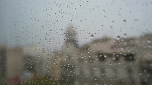 A slow-motion footage of raindrops seen through a window during heavy rainfall, with an urban city l
