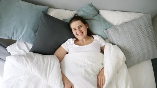 Young woman lying in bed under blanket, opens it and smiles in camera