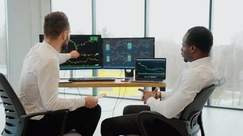 Two Diverse Colleagues Traders Talking to Each Other Looking at Graphs While Sitting in the Office