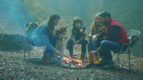 Family Sit Around a Campfire on Autumn Evening in the Woods Frying Sausages Over a Fire in Forest