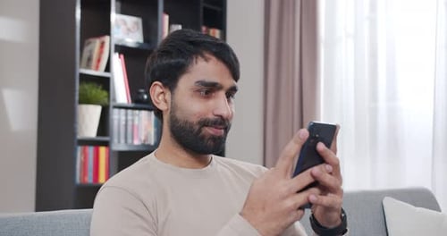 Close Up of Happy Arab Young Man Sitting on Sofa and Chatting By Messages on Smartphone At Home