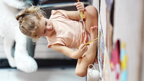 Little Girl Sits on the Floor at Home and Draws with Paints and Brushes in a Coloring Book
