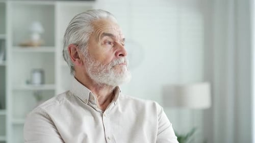 Close up portrait of a serious elderly man with gray hair sitting on a sofa in a bright room at home