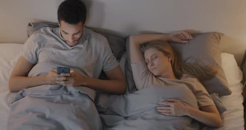 Hombre afroamericano que envía mensajes de texto con teléfonos inteligentes y mira a su esposa dormida, mujer caucásica en