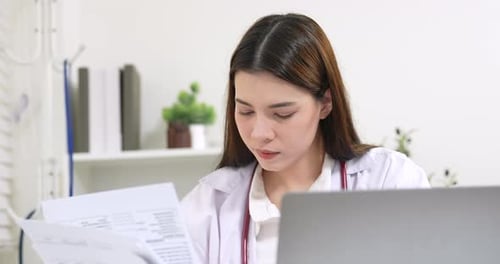 Asian female pediatrician reading medical documents beside laptop, serious expression, showing