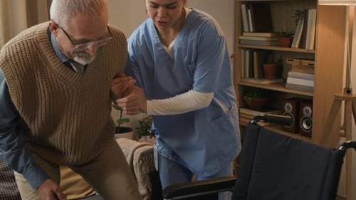 Caring Nurse Helps Senior Man Into Wheelchair Indoors