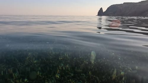 Sea Water Surface Camera Flies Over the Calm Azure Sea with Volcanic Rocky Shores on Background
