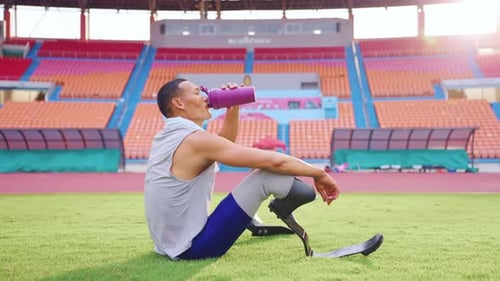 Asian para-athlete with prosthetic blades drinking water in stadium.