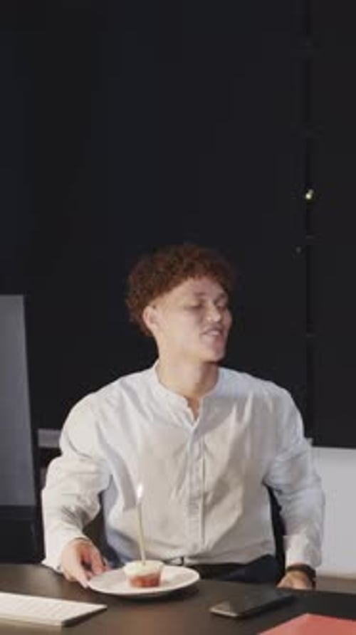 Vertical video: Diverse coworkers serving birthday cupcake, blowing candle and eating treat at desk
