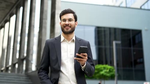 Confident businessman in a formal suit uses mobile phone while walking on the street near modern