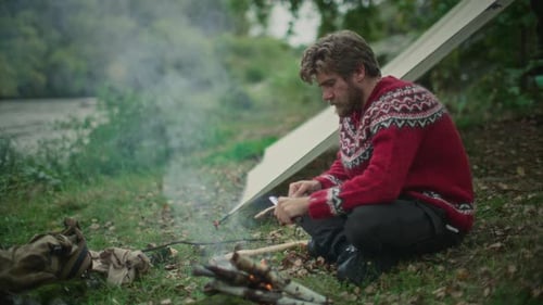 Man Carving Wooden Stick beside Campfire and Tarp Shelter in Forest
