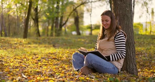 Adult Woman Sitting Under Tree Reading a Book in Autumn Park