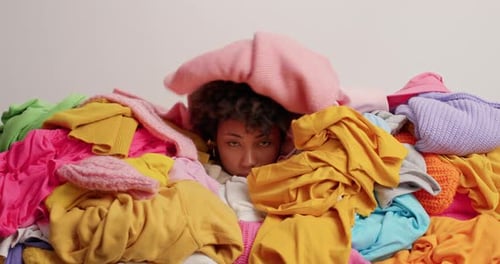 Sad young woman drowned in laundry heap of clothes against white background