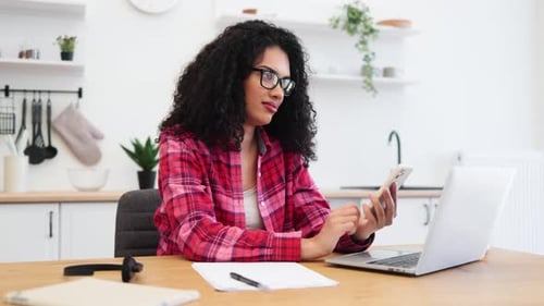 Young Woman Using Smartphone at Home While Working Remotely on a Laptop