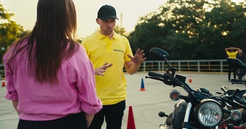 Un instructor de conducción con una camiseta amarilla le dice a una estudiante con una camiseta rosa cómo comportarse mientras
