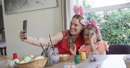 Mom and daughter taking selfie with mom extending phone while painting eggs at table for Easter