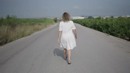 Young Curly Haired Blonde Caucasian Woman in Short White Dress Walking with Her Back to Camera on