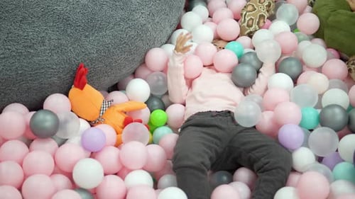 Cheerful little girl plays in a dry pool lying in white, pink and grey plastic balls